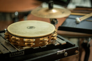 A close-up of percussion instruments, prominently featuring a tambourine placed on top of a Deagan xylophone, with drumsticks resting on a music stand and a cymbal in the background.