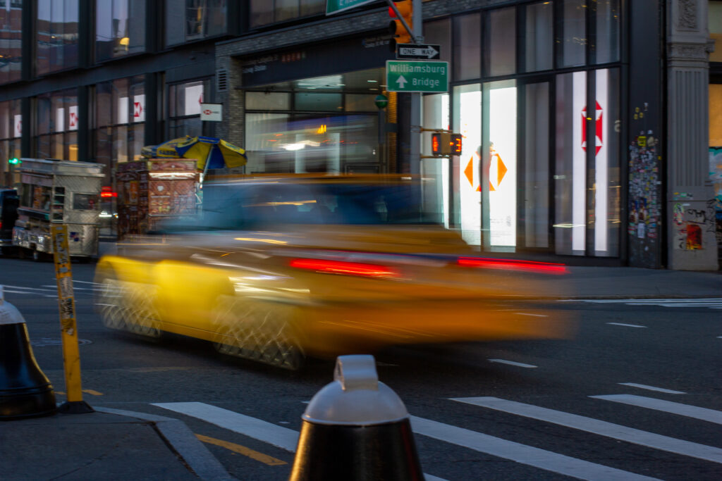 A yellow cab moves quickly down a New York City street with food carts and a bank in the background.