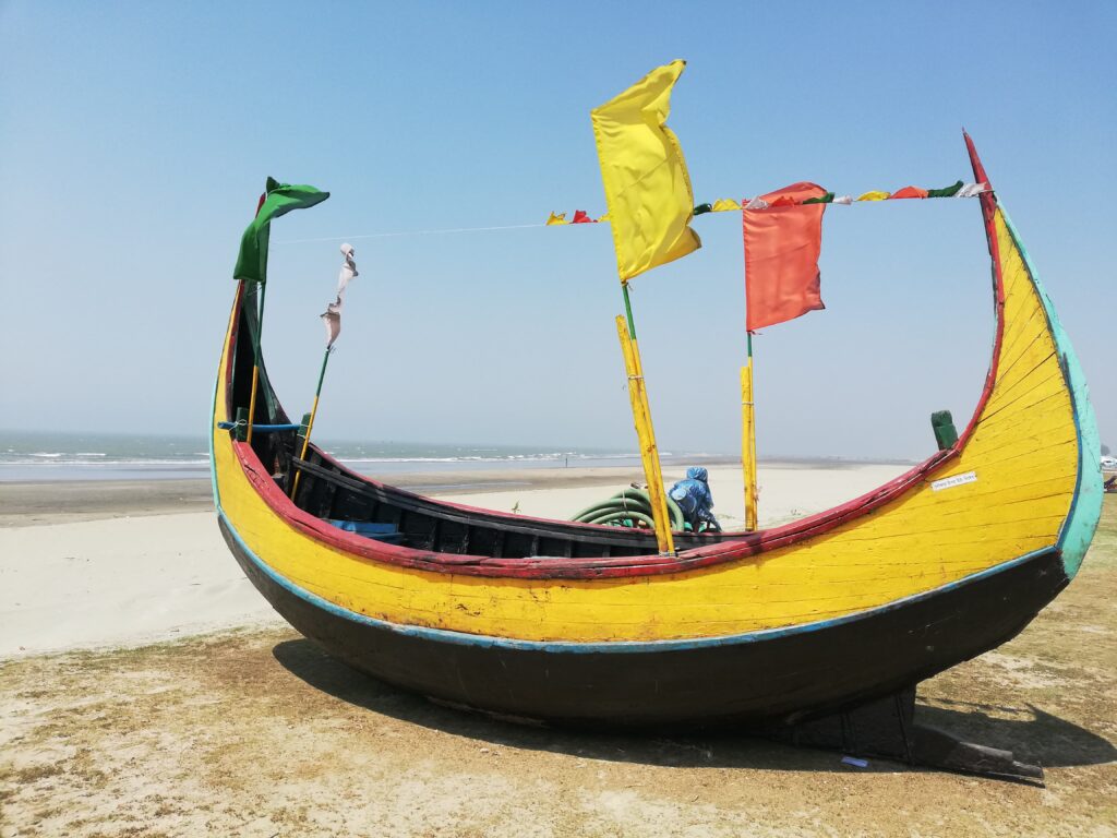 A colorful wooden boat with a high curved prow decorated with flags moored on a sandy beach with the ocean in the background on a clear day.