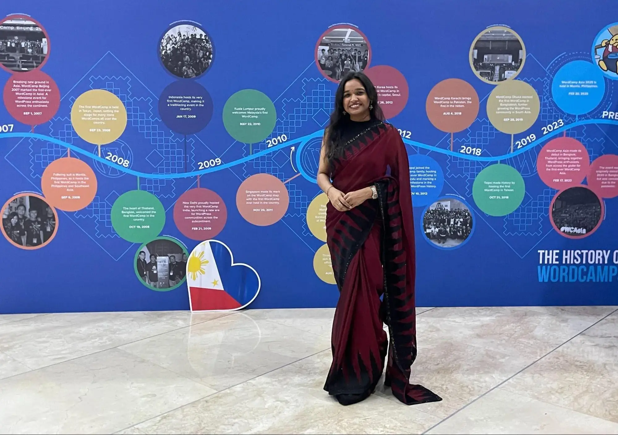 Astha in a long dark sari, standing in front of a wall with a timeline of WordPress on it, at WordCamp Asia in Philippines.
