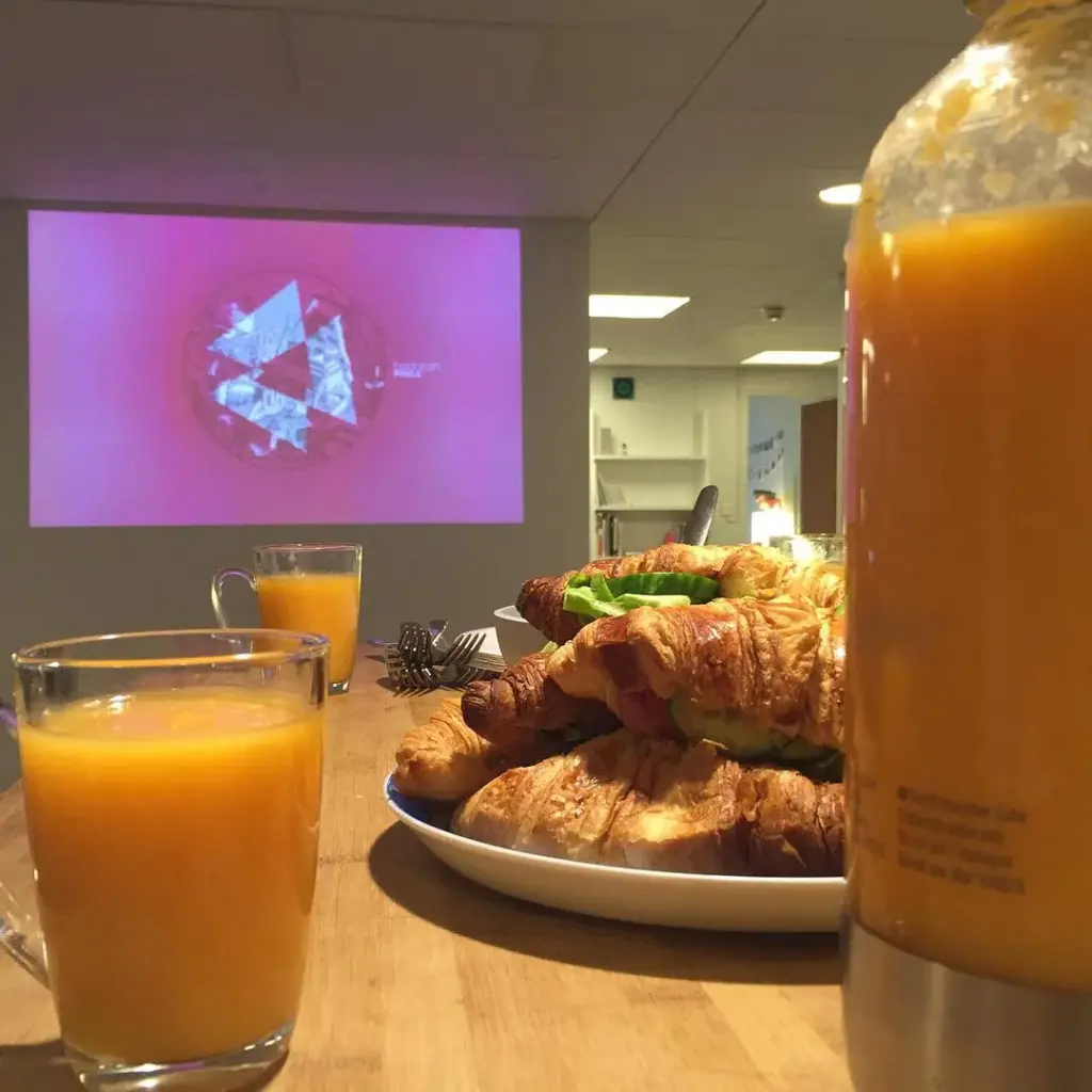 Closeup of a glass of orange juice with croissants next to it, and a projection screen in the background.